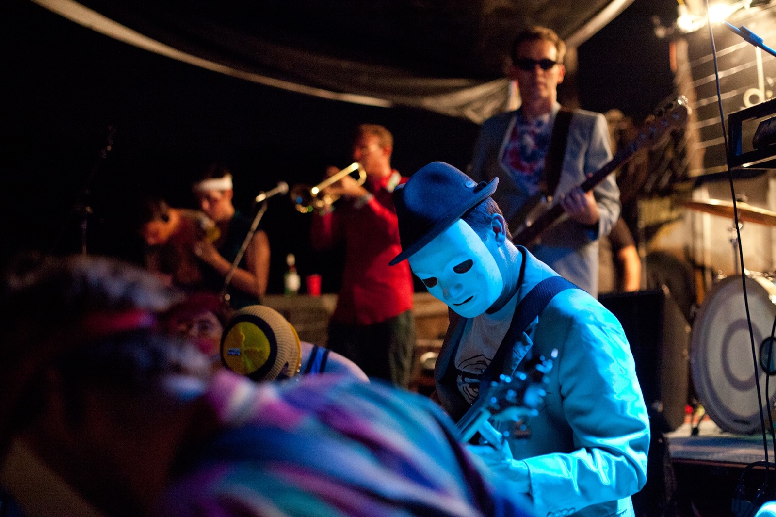 Masked guitarist in a bowler hat under blue light with full band